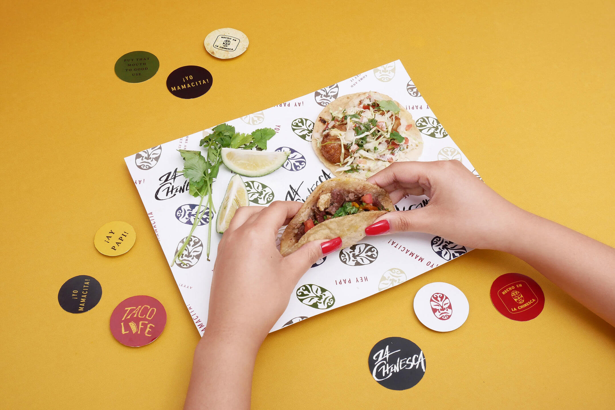 Hands holding a soft shell La Chinesca taco surrounded by branded stickers
